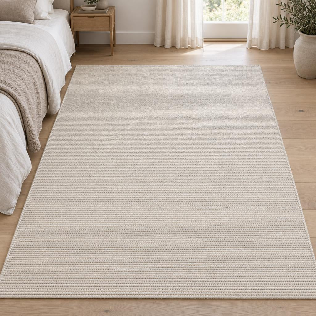 Vloerkleed Copenhagen Crème, vlakgeweven wol met katoen in grove ribbels, naast een linnen bed – sfeerimpressie in de slaapkamer