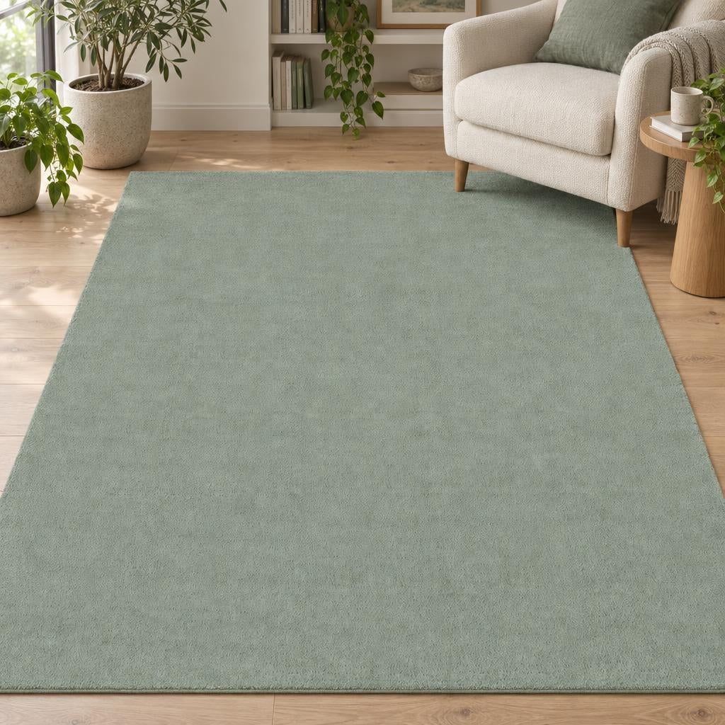 Effen vloerkleed Cozy Groen in zacht saliegroen, laagpolig, met een crème fauteuil en planten – sfeerimpressie in de woonkamer