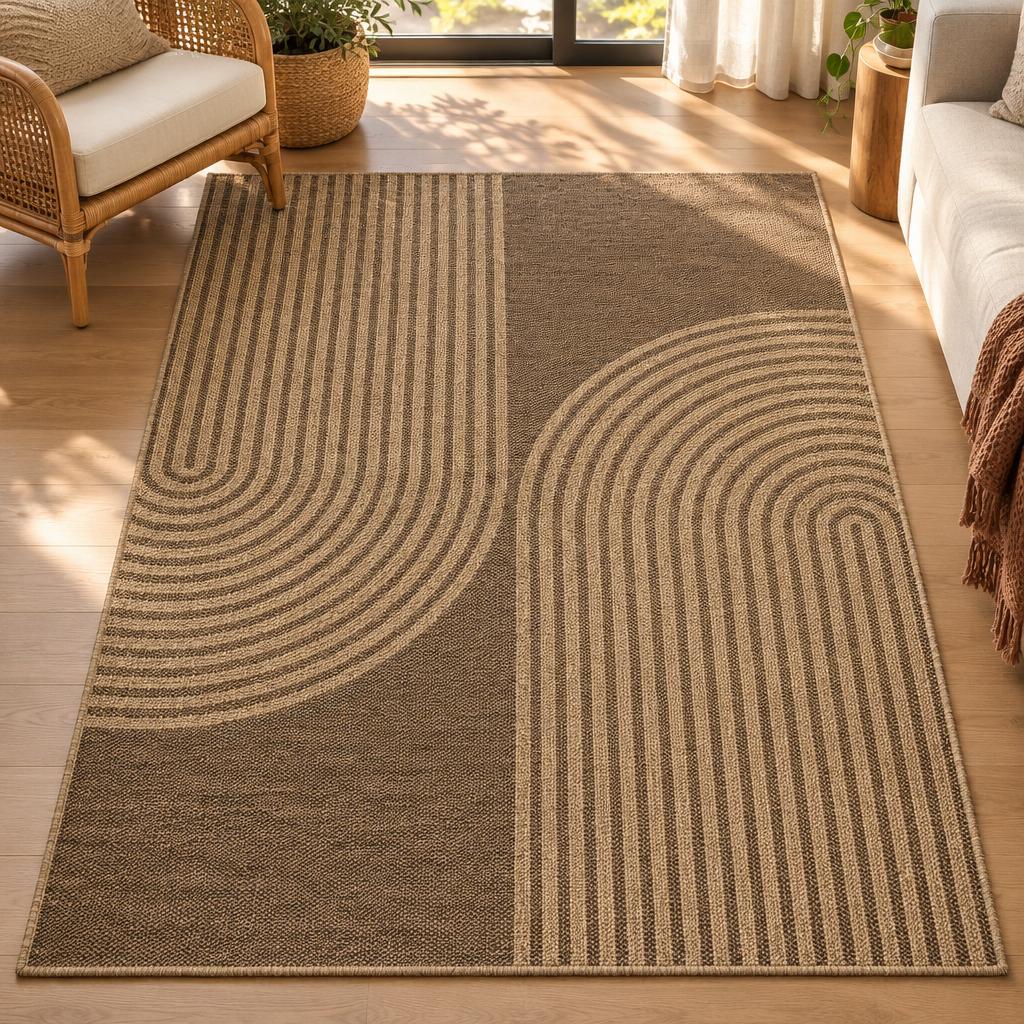 Vloerkleed Kenzy 5029 Naturel, vlakgeweven in koffiebruin met zandkleurige boogstrepen, naast een rotan fauteuil – sfeerimpressie in de woonkamer